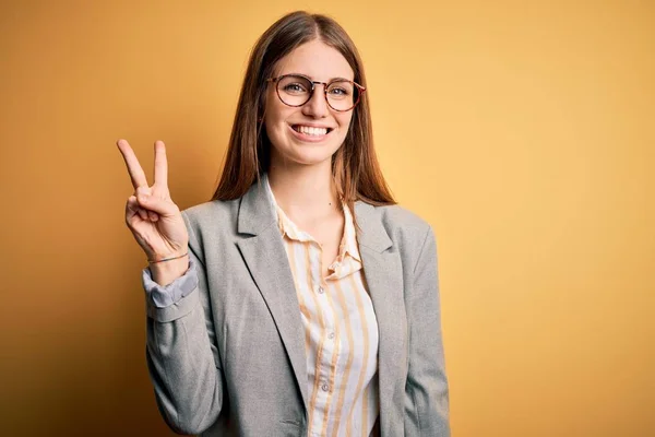 年轻美丽的红头发女人 穿着夹克和眼镜 站在孤立的黄色背景上 带着自信和快乐的笑容 用手指指着二号 — 图库照片