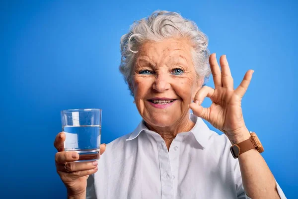年长美丽的女人站在孤零零的蓝色背景上喝着一杯水 手拿着手签 是极好的象征 — 图库照片