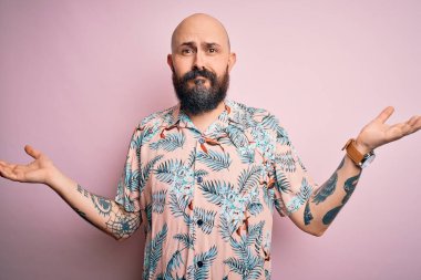 Sakallı ve dövmeli yakışıklı, pembe arka planda sıradan bir çiçek gömleği giyen, elleri ve kolları havada, kafası karışık bir adam. Şüphe kavramı.
