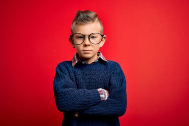 Mavi gözlü küçük beyaz çocuk kırmızı arka planda gözlük takıyor şüpheci ve gergin, yüzünde çapraz kollu tasvip etmeyen bir ifade. Negatif kişi.