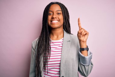 Genç Afro-Amerikan iş kadını pembe izole edilmiş arka planda duruyor ve bir numaralı parmağını gösterirken kendinden emin ve mutlu bir şekilde gülümsüyor..