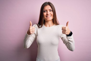 Mavi gözlü genç bir kadın pembe arka plan işareti üzerine gündelik beyaz tişört giyip el hareketleriyle olumlu jest yapıyor, gülümsüyor ve mutlu. Neşeli ifade ve kazanan jest.