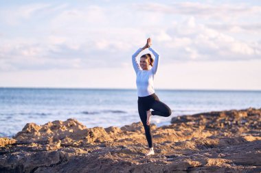 Genç ve güzel sporcu gülümseyerek yoga yapıyor. Yüzünde gülümseme olan koç sahilde ağaçlara poz veriyor.
