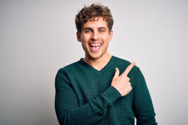 Sarışın, kıvırcık saçlı, yeşil süveter giyen, beyaz arka planda neşeli yüzlü, elleri ve parmaklarıyla gülümseyen, yüzünde mutlu ve doğal bir ifade olan genç bir adam.