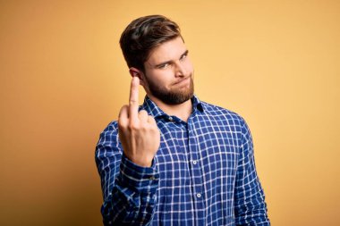 Sarışın, sakallı ve mavi gözlü genç bir işadamı sarı arka planda tişört giyiyor orta parmağını gösteriyor, kaba ve kaba bir ifade sergiliyor.