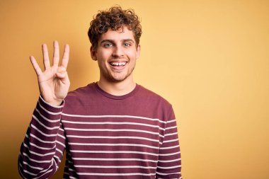 Sarışın, kıvırcık saçlı, sıradan çizgili süveter giyen, dört numarayı gösteren, kendinden emin ve mutlu gülümseyen genç bir adam..