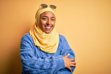 Sarı Müslüman başörtüsü ve güneş gözlüğü takan, kollarını kavuşturup kameraya gülümseyen genç Afrikalı Amerikalı öğrenci. Pozitif kişi.