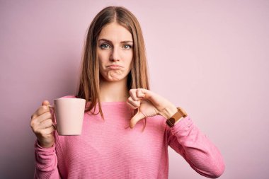Genç, güzel, kızıl saçlı bir kadın izole edilmiş pembe arka planda kahve içiyor. Kızgın bir yüzü var. Negatif işaretleri var.