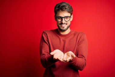 Sakallı, gözlüklü, süveterli, kırmızı arka planda duran, elleri avuçlarında gülümseyen ve el hareketi yapan yakışıklı bir adam. Tutun ve koruyun