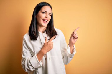 Genç esmer, mavi gözlü spor kadın sarı arka planda eşofman üstü giyiyor gülümsüyor ve kameraya bakıyor iki eliyle ve parmaklarıyla yandan işaret ediyor..