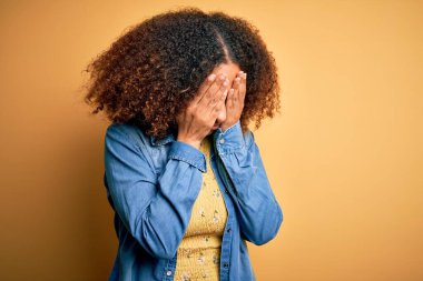 Afro saçlı, sarı arka planda sıradan kot gömlek giyen, elleri ve elleri ile yüzünü kapatan, ağlayan Afro-Amerikalı genç bir kadın. Depresyon kavramı.