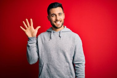 Eşofman giyen genç yakışıklı sporcu izole edilmiş kırmızı arka plan üzerinde duruyor ve beş numaralı parmaklarıyla işaret ederken kendinden emin ve mutlu bir şekilde gülümsüyor..