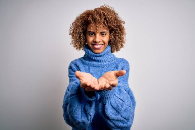 Beyaz arka planda boğazlı kazak giyen, elleri avuçlarında gülümseyen ve jest yapan güzel Afro-Amerikan bir kadın. Tutun ve koruyun