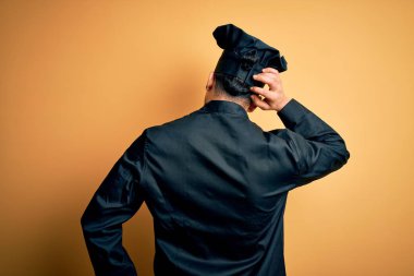 Aşçı üniforması giyen genç Brezilyalı aşçı ve izole edilmiş sarı arka planda şapkası var. Geriye doğru, elleri kafasında şüpheyi düşünüyor.
