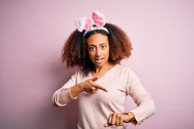 Afro saçlı, pembe arka planda tavşan kulağı takan Afro-Amerikalı genç bir kadın. Aceleyle saate, sabırsızlığa, gecikmeye kızgın ve sinirli bakıyor.