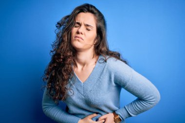 Kıvırcık saçlı genç bir kadın izole edilmiş arka planda mavi bir süveter giyiyor. Midesinde hazımsızlık, acı verici bir hastalık var. Ache kavramı.