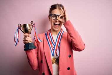 Genç, güzel, kızıl saçlı bir kadın pembe arka planda madalyalar takıyor, gülümsüyor ve el pençe divan duruyor.