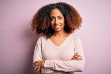 Afro saçlı, pembe arka planda rahat süveter giyen, kollarını kavuşturup kameraya gülümseyen Afro-Amerikan genç bir kadın. Pozitif kişi.