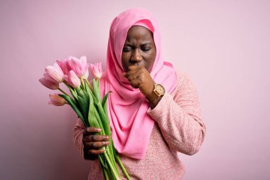 Genç Afro-Amerikan, Müslüman başörtüsü takmış, elinde bir buket pembe lale tutan ve soğuk algınlığı veya bronşitin belirtisi olarak öksüren iri yarı bir kadın. Sağlık hizmeti kavramı.