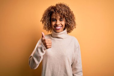 Genç ve güzel Afro-Amerikan kadın balıkçı yaka kazak giyip sarı arka planda mutlu bir el hareketi yapıyor. Kameraya bakıp başarıyı gösteren ifade onaylanıyor.