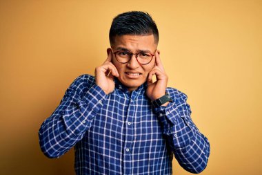 Sarı arka plan üzerine gündelik gözlük takan genç, yakışıklı latin adam yüksek sesli müzik sesi için kulaklarını parmaklarıyla kapatıyor. Sağır kavramı.