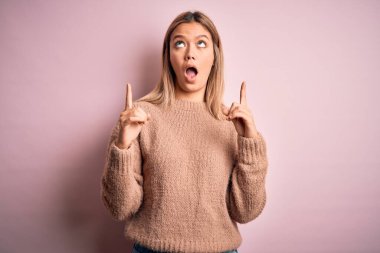 Pembe izole arka planda, kışlık yün kazak giyen genç, güzel sarışın kadın şaşırmış ve şaşırmış bir şekilde yukarı bakıyor ve parmaklarıyla işaret ediyor ve kollarını kaldırıyordu..