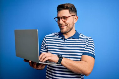 Mavi gözlü genç bir iş adamı bilgisayar bilgisayarı kullanıyor. Mutlu bir yüzü var. Dişlerini gösteren kendine güvenen bir gülümsemeyle ayakta duruyor.