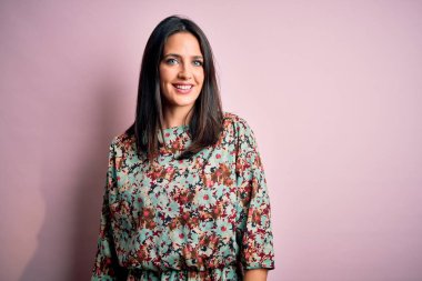 Mavi gözlü genç esmer kadın pembe arka planda çiçekli elbiseler giyiyor ve yüzünde mutlu ve havalı bir gülümseme var. Şanslı kişi..