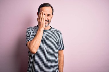 Orta yaşlı, beyaz bir adam günlük çizgili tişört giyiyor. İzole edilmiş pembe arka planda duruyor. Bir gözünü eliyle kapatıyor..