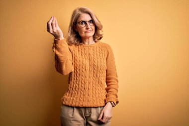 Orta yaşlı güzel sarışın kadın sarı arka planda günlük süveter ve gözlük takıyor. İtalyan jestleri yapıyor. Elleri ve parmakları kendinden emin.