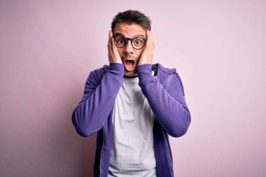 Pembe arka planda duran mor kazak ve gözlük takan genç yakışıklı adam korkmuş ve şok olmuş, şaşırmış ve şaşırmış bir halde.