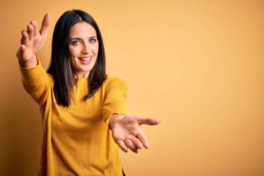 Mavi gözlü genç esmer kadın sarı arka planda sıradan bir süveter giyiyor ve kameraya sarılıp gülümsüyor. Mutluluğu kucaklayan neşeli bir ifade..