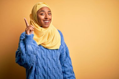 Genç Afrikalı Afro kadın, izole edilmiş sarı arka planda Müslüman başörtüsü takıyor. Bir numarayı işaret ediyor ve gülümsüyor. Kendinden emin ve mutlu..