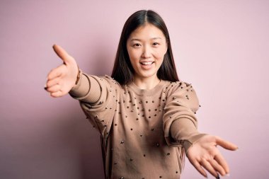 Genç, güzel Asyalı bir kadın şık bir süveter giyerek pembe arka planda kameraya bakıp sarılmak için kollarını açmış gülümsüyor. Mutluluğu kucaklayan neşeli bir ifade..