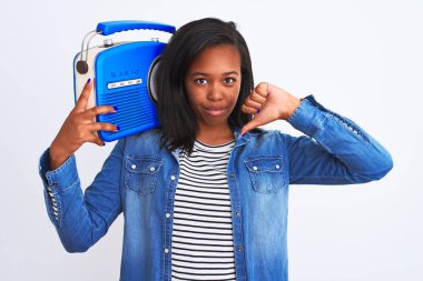 Genç Afrikalı Amerikalı bir kadın, izole edilmiş arka planda antika bir radyo tutuyor. Kızgın bir yüzü var. Negatif işaretler, baş parmakları aşağıda, reddedilme kavramından hoşlanmadığını gösteriyor.