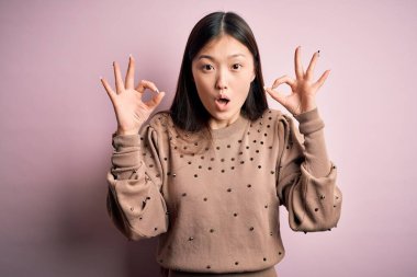 Pembe arka planda şık süveter giyen genç ve güzel Asyalı bir kadın parmaklarıyla onay sembolü yaparken şaşırmış ve şok olmuş görünüyor. Çılgın ifade
