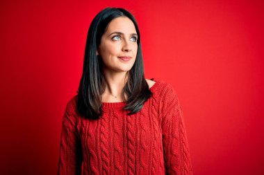 Mavi gözlü genç esmer kadın izole edilmiş kırmızı arka planda günlük süveter giyiyor. Gülümsüyor ve uzaklara bakıyor..