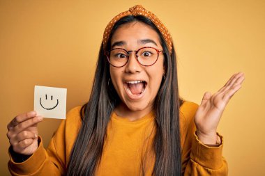 Asyalı kadın, elinde gözlüklü, gülümseyen emojiyle sarı arka planda mutlu ve heyecanlı bir ifade çiziyor. Kazanan ifade zaferi büyük bir gülümsemeyle kutluyor ve ellerini kaldırıyor.