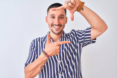 Deniz çizgili gömlek giyen yakışıklı genç adam izole edilmiş arka planda duruyor gülümsüyor ve elleri ve parmakları gülen yüzlü bir çerçeve yapıyor. Yaratıcılık ve fotoğrafçılık kavramı.