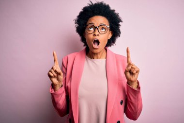 Kıvırcık saçlı, pembe ceketli, güzel Afro-Amerikan iş kadını şaşırmış ve şaşırmış bir şekilde yukarı bakıyor ve parmaklarıyla işaret ediyor ve kollarını kaldırıyor..