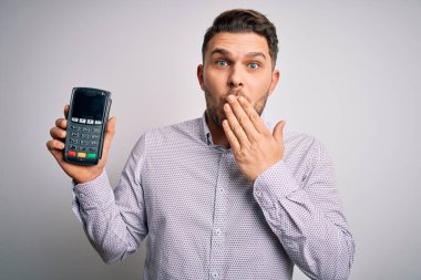 Young business man with blue eyes holding dataphone payment terminal over isolated background cover mouth with hand shocked with shame for mistake, expression of fear, scared in silence, secret concept clipart