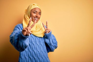 İzole edilmiş sarı arka plan üzerinde Müslüman başörtüsü takan genç Afro kadın kameraya bakıp zafer işareti yaparken gülümsüyor. İki numara..