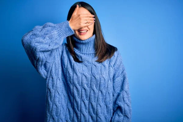 Young brunette woman with blue eyes wearing casual turtleneck sweater smiling and laughing with hand on face covering eyes for surprise. Blind concept.