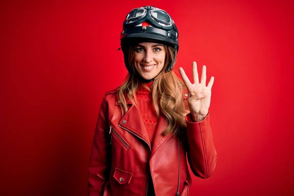 若いです美しいですブルネットMotoヘルメットを身に着けています上の赤い背景表示と指差しますとともに指番号4ながら笑顔自信と幸せ — ストック写真