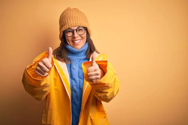 Middle age woman wearing yellow raincoat and winter hat over isolated background approving doing positive gesture with hand, thumbs up smiling and happy for success. Winner gesture.