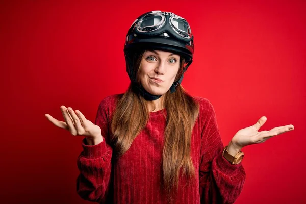 赤い背景の上に元ヘルメットを身に着けている若い美しい赤毛の元サイクリストの女性は 腕や手を上げていると混乱した表情 疑いの概念 — ストック写真