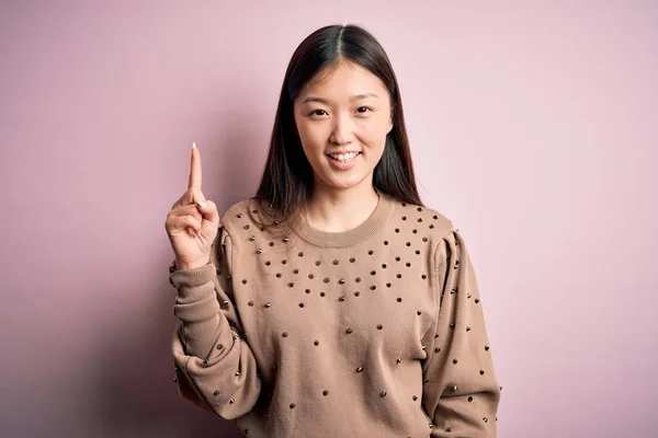 年轻美丽的亚洲女人穿着时尚和雅致的毛衣 穿着粉色的舒适的背景 手指指向成功的想法 又兴奋又快乐第一部分 — 图库照片