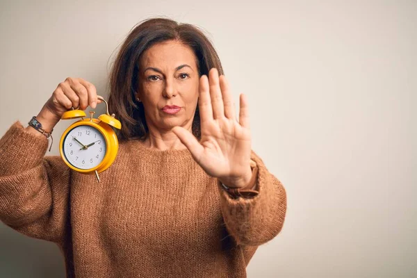 孤立した背景の上に古典的な目覚まし時計を保持している中世のブルネットの女性は手の手のひらで歌うのを止める 顔に負と深刻なジェスチャーで警告式 — ストック写真