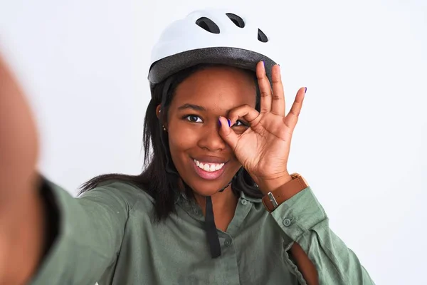 Young african american woman wearing bike helmet taking a selfie over isolated background with happy face smiling doing ok sign with hand on eye looking through fingers