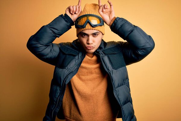 Молодой бразильский лыжник в снежной спортивной одежде и лыжных очках на желтом фоне делает смешной жест с пальцем над головой, как рога быка
